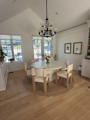 A dining area with a round table, chairs, a chandelier, and a view through large windows.