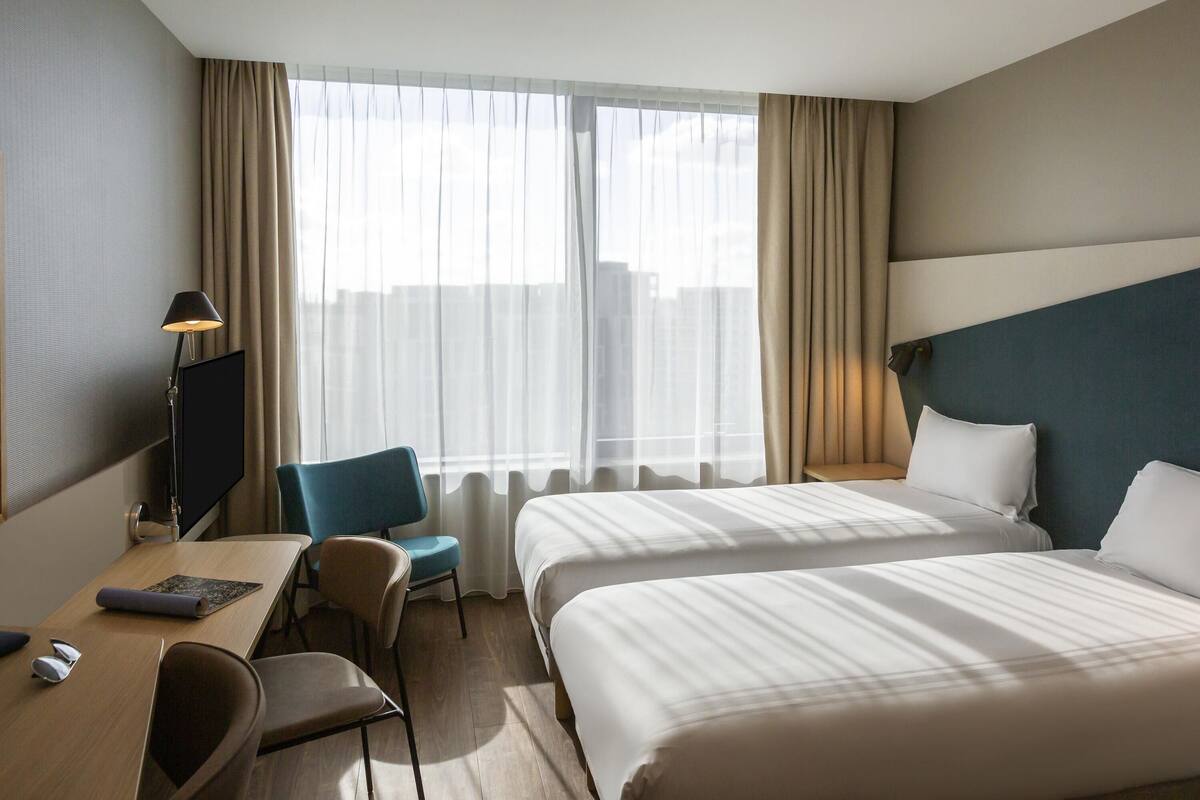 Une chambre d’hôtel avec deux lits, un bureau avec une chaise, une télévision et une fenêtre avec des rideaux.