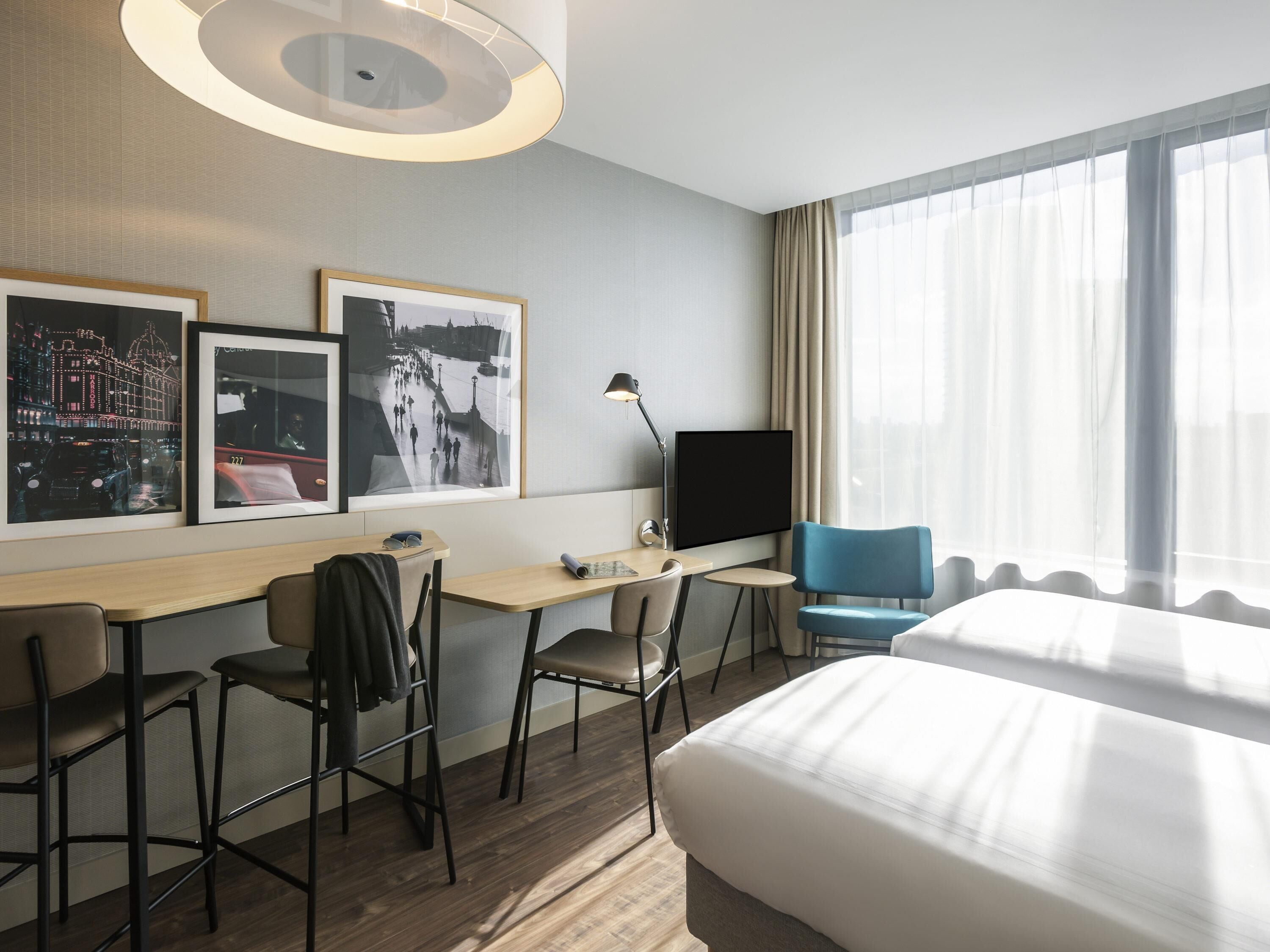 A hotel room with a bed, desk, chairs, a TV, and large windows.