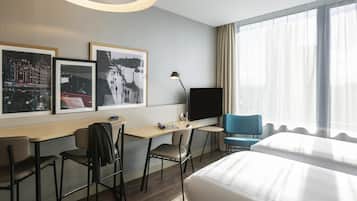 A hotel room with a bed, desk, chairs, a TV, and large windows.