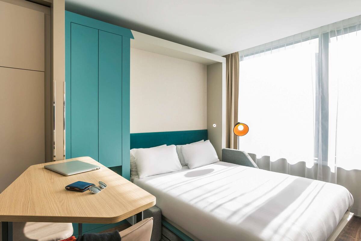 Une chambre d’hôtel comprenant un lit, un bureau, une chaise, une armoire et une fenêtre avec des rideaux.