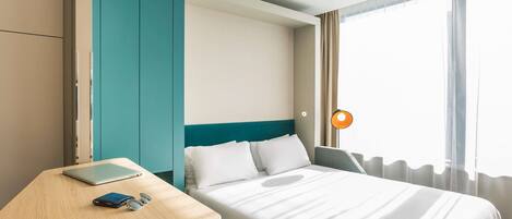 A hotel room with a bed, a desk, a chair, a wardrobe, and a window with curtains.