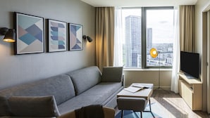 A modern hotel room with a gray sofa, a small table with a book, a TV, and framed artwork on the wall. - Aparthotel Adagio London Stratford (London)