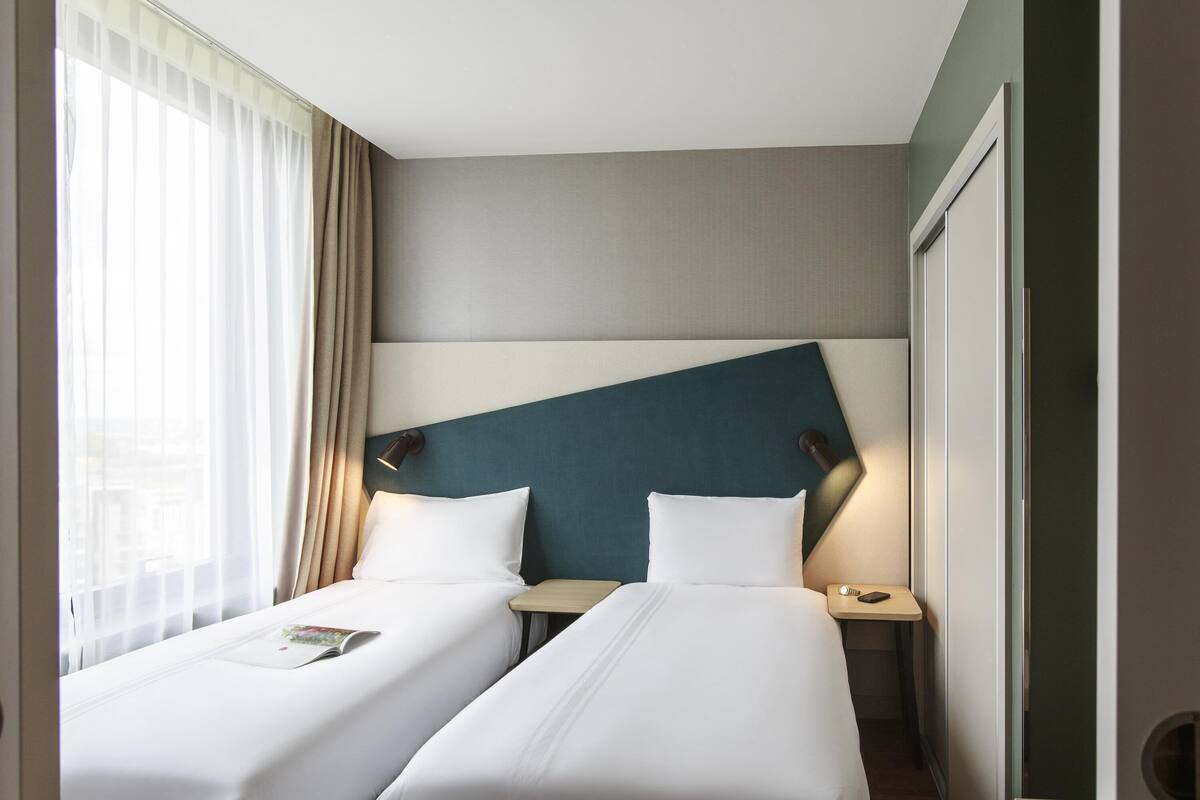 Une chambre d’hôtel avec deux lits simples, une table de chevet avec une lampe, une fenêtre avec des rideaux légers et une vue sur la ville.