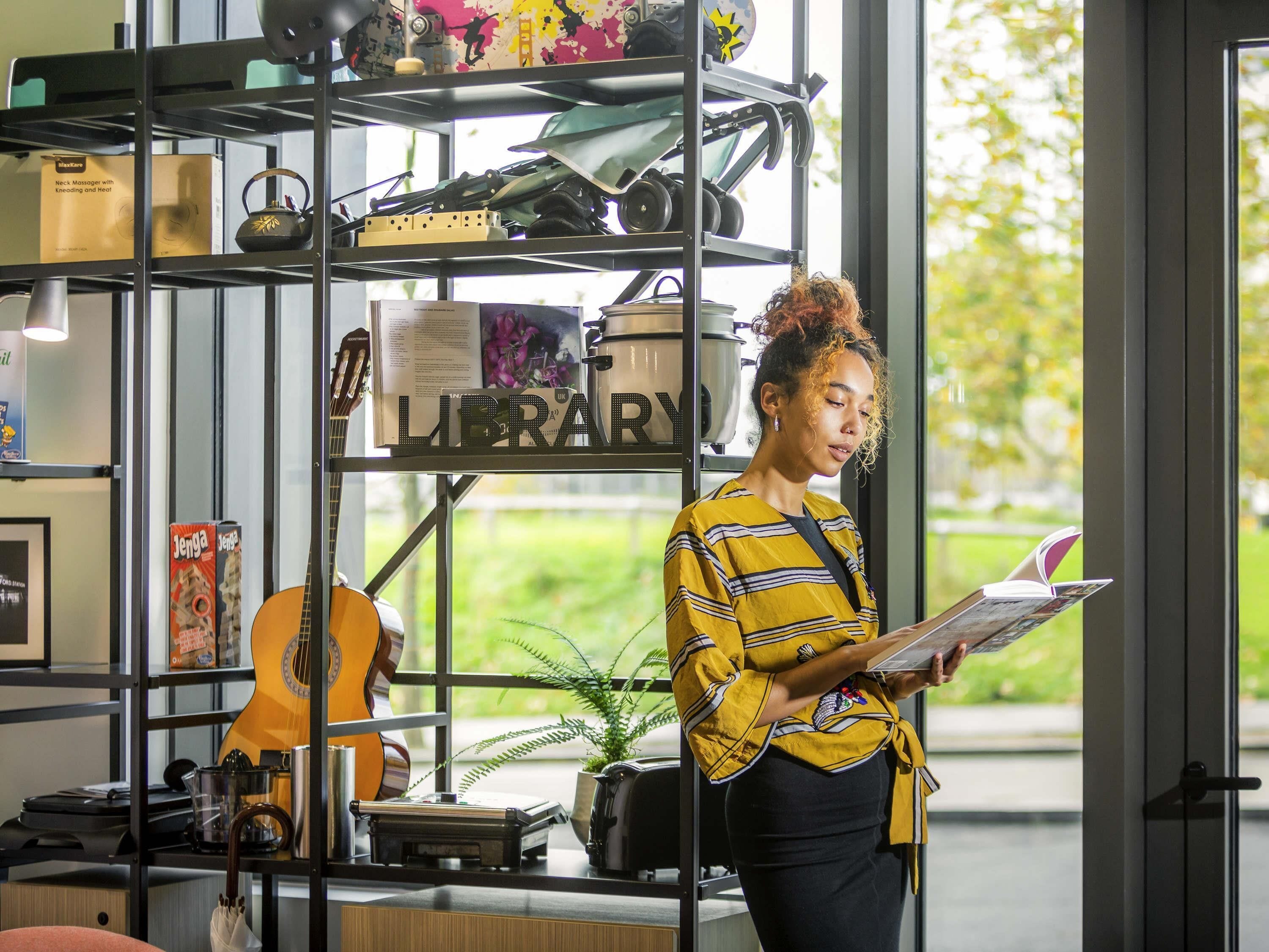 A person reading a book in a room with a bookshelf containing various items, a guitar, and a plant.
