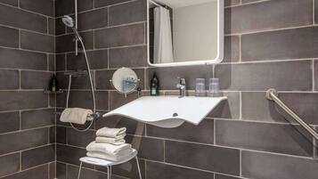 Een moderne badkamer met donkere tegels, een witte wastafel, een ronde spiegel, een douche en een handdoekrek.