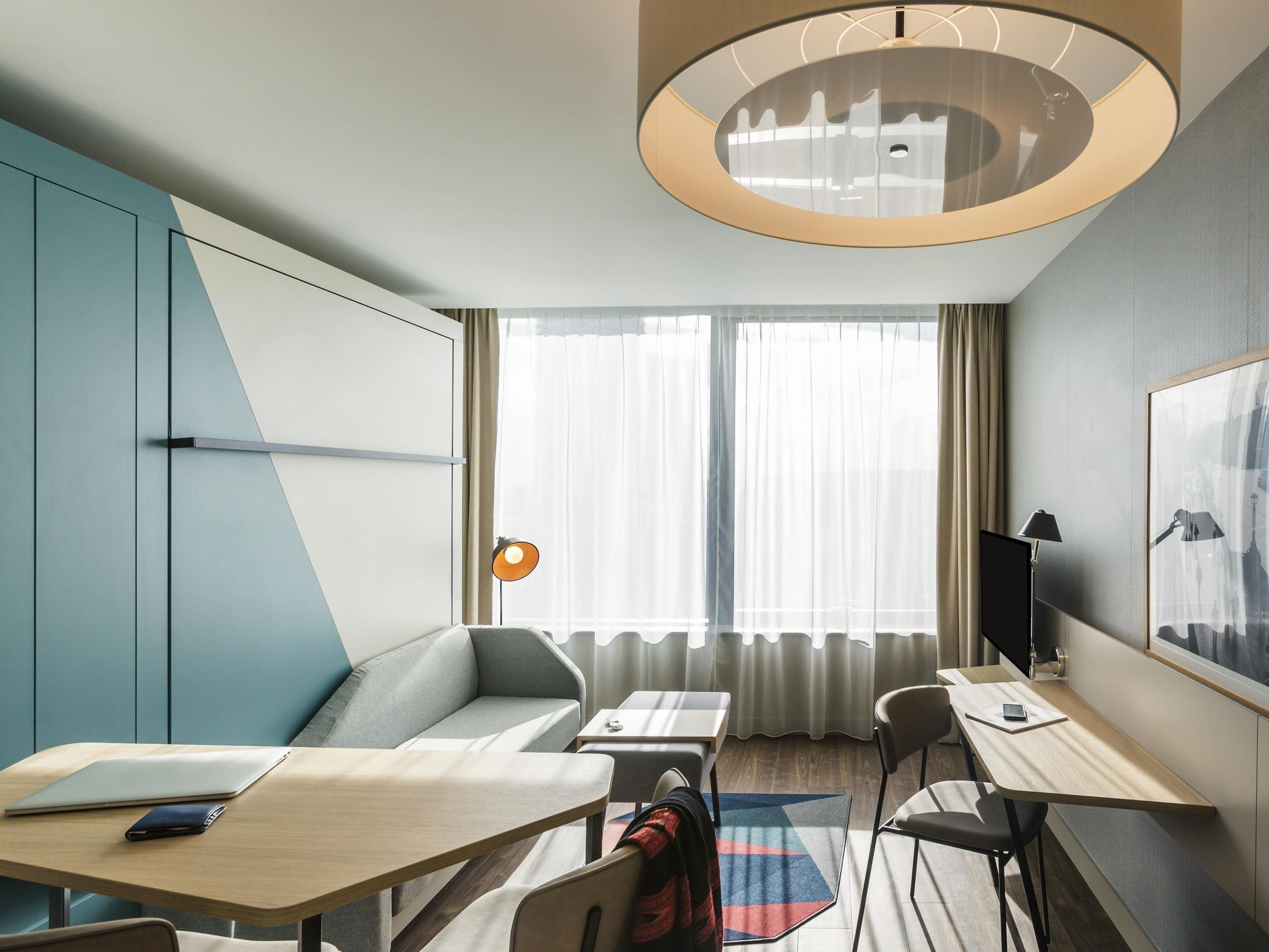 A modern hotel room with a sofa, a desk, a chair, a laptop, a mobile phone, a lamp, and a large window with curtains.