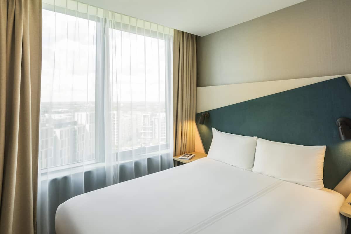 Une chambre d’hôtel avec un grand lit, une vue sur la ville par la fenêtre et une table de chevet avec une lampe.