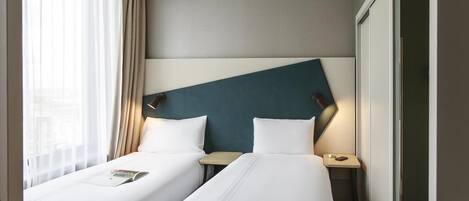 Ein Hotelzimmer mit zwei Einzelbetten, einem Nachttisch mit Lampe und einem Fenster mit Vorhängen.