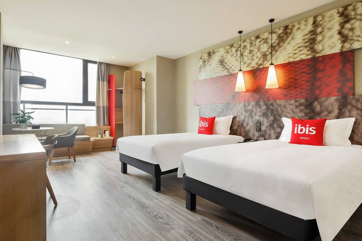 A hotel room with two beds, a desk, a chair, a sofa, a window, and a plant.