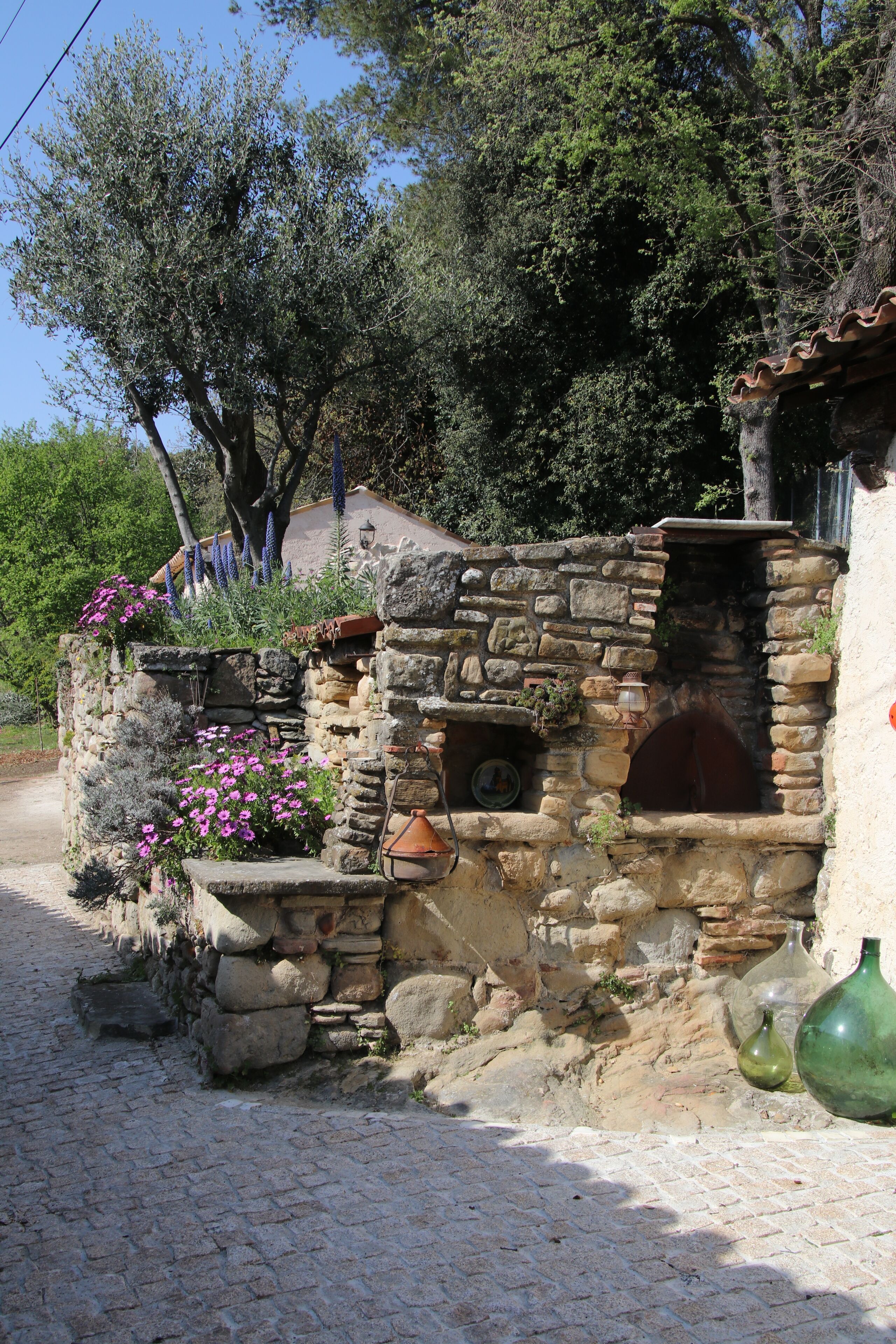 A rustic stone building with a brick oven, surrounded by flowering plants and trees.