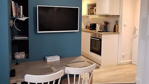 A modern kitchen with white cabinets and a microwave, a dining area with a wooden table and white chairs, and a bookshelf with books and a clock. - "Blue Dream" – Cozy, clean & stylish studio | Doua / INSA / CESI - Garage, WIFI (Villeurbanne)