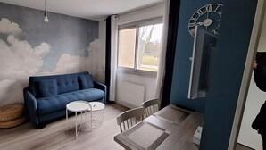 A room with a blue sofa, a small round table, and a dining table with chairs. There is a window with a view of the outside, and a wall clock. - "Blue Dream" – Cozy, clean & stylish studio | Doua / INSA / CESI - Garage, WIFI (Villeurbanne)