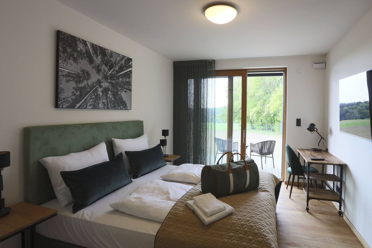Una habitación de hotel con cama, escritorio, silla, ventana con vistas a árboles y un cuadro en la pared.
