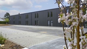 A modern building with a flat roof, large windows, and a paved area in front.