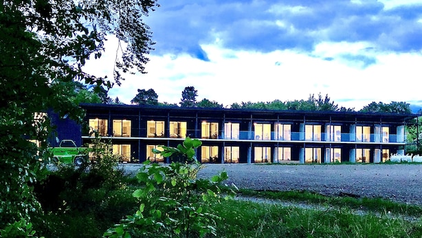 A long, modern building with multiple windows and a flat roof, surrounded by greenery.