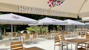 An outdoor seating area with white tables and chairs under large umbrellas, featuring a logo with a red and black crest.