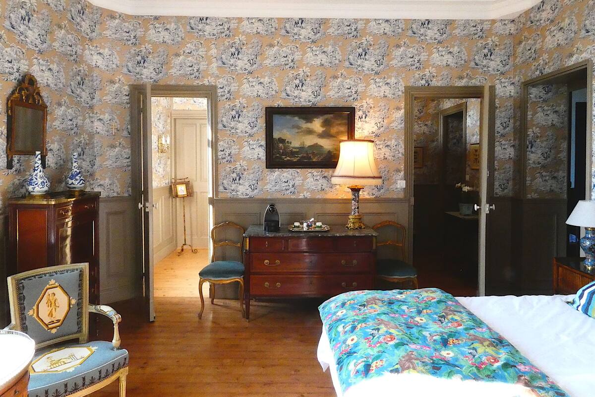 A room with floral wallpaper, a bed with a floral bedspread, a dresser, a chair, and a lamp.