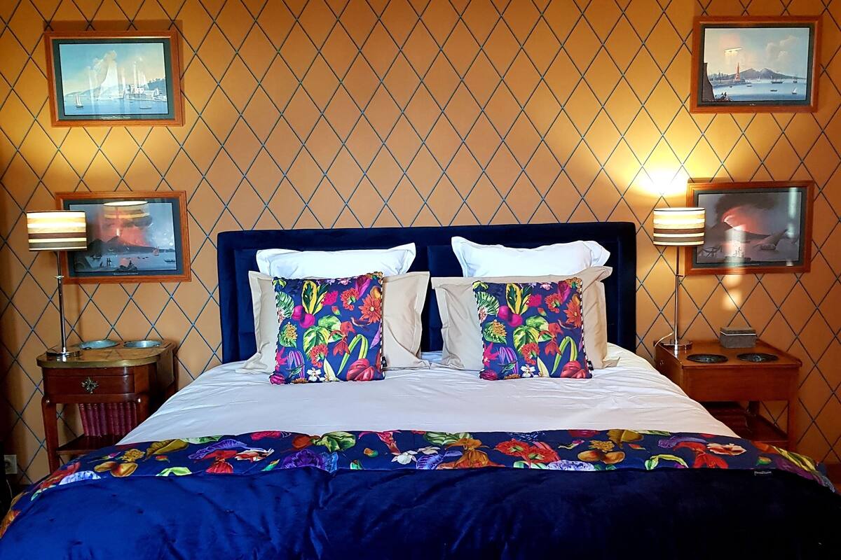 A bed with a blue bedspread and floral pillows, flanked by two nightstands with lamps and framed pictures on the wall.