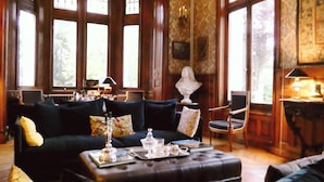A spacious living room with dark blue sofas, a coffee table with tea set, and a chandelier. Large windows allow natural light to fill the room. - Chateau de Hieville (Saint-Pierre-en-Auge)