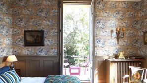 A bedroom with a bed, a chair, a table with a lamp, a painting, and a view of the outdoors. - Chateau de Hieville (Saint-Pierre-en-Auge)