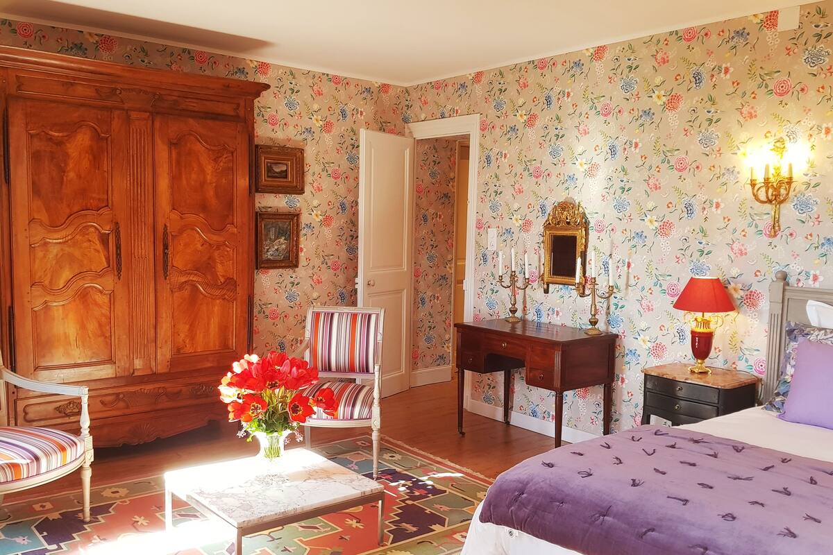 A bedroom with a large wooden wardrobe, a striped chair, a desk with a mirror, a bedside table with a lamp, and a bed with a purple cover.