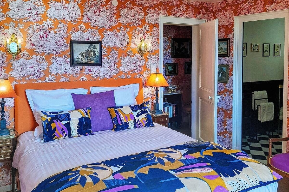 A bedroom with a floral wallpaper, a bed with colorful bedding, two bedside lamps, and a door leading to a bathroom.