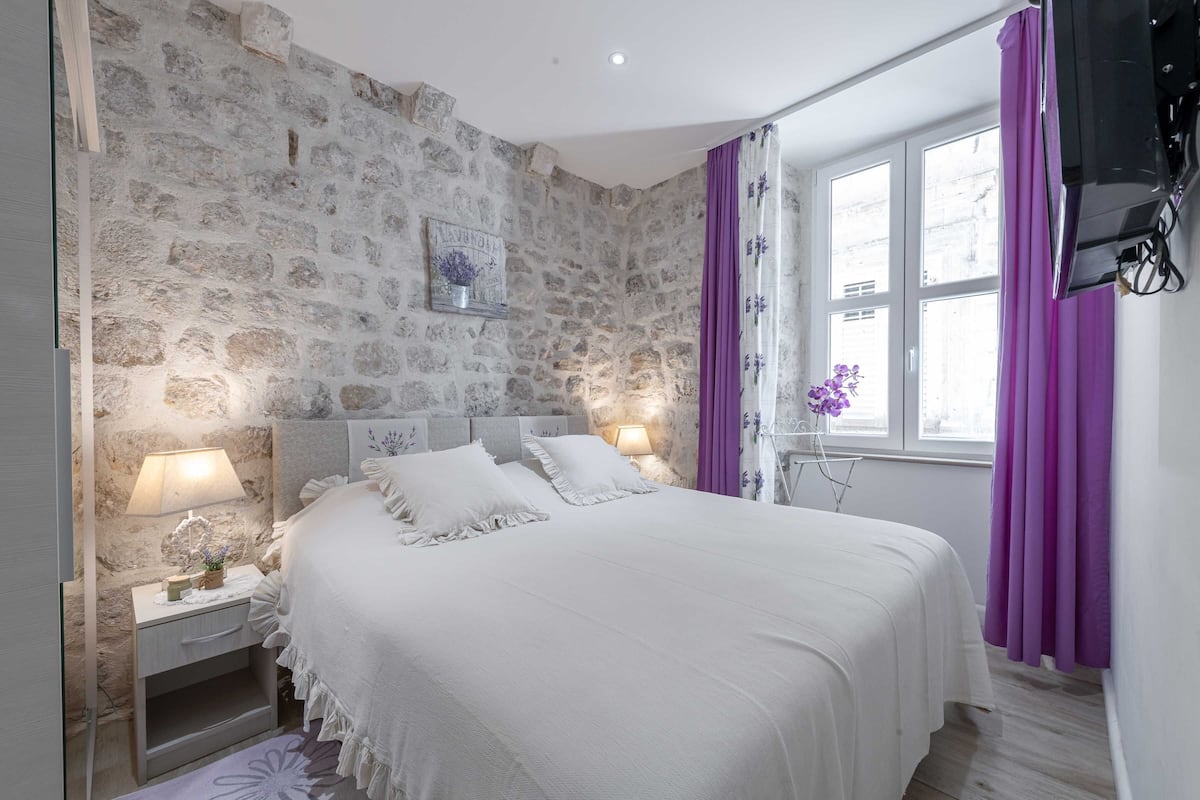 A bedroom with a stone wall, a bed with white bedding, purple curtains, and a window with a view.