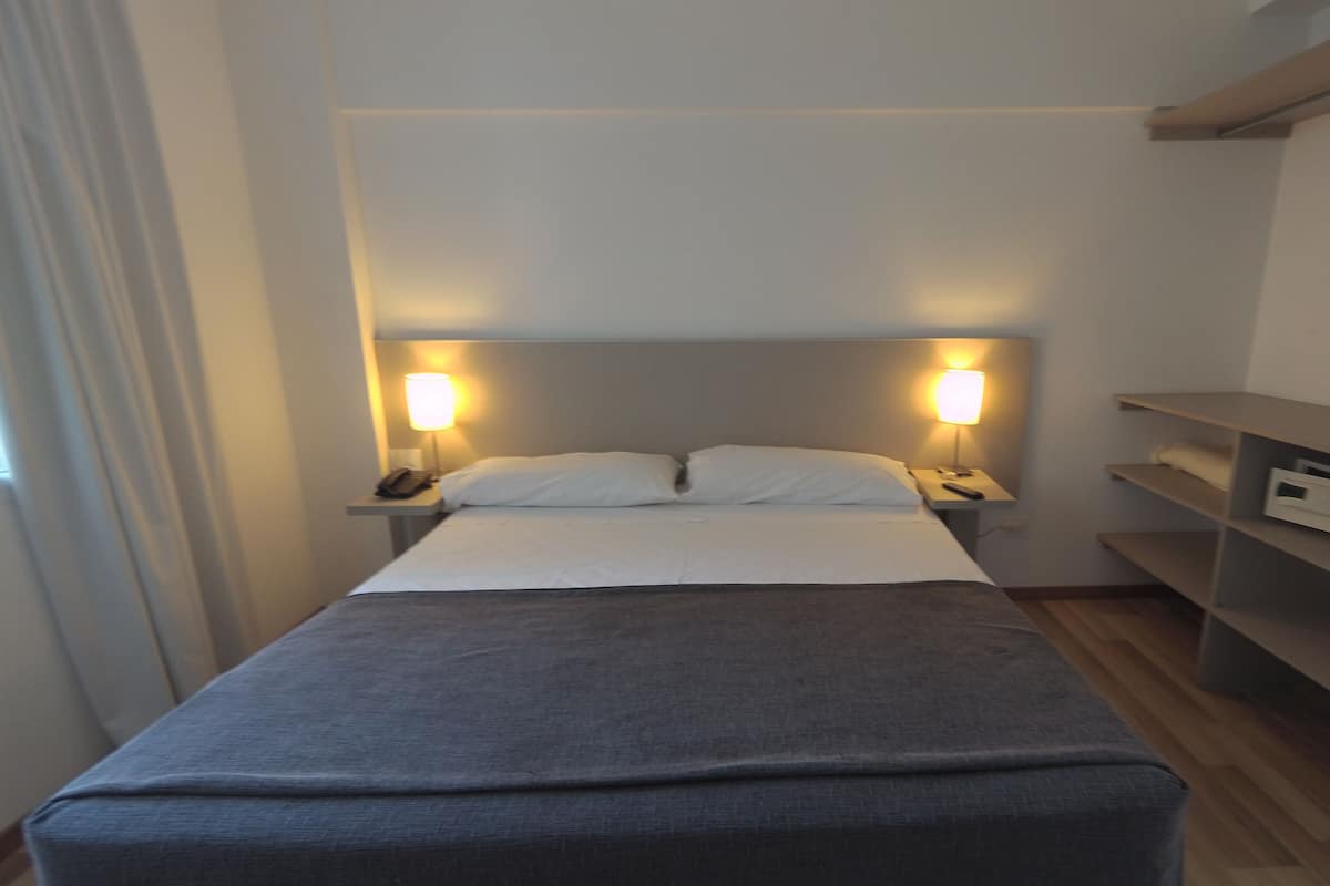 A neatly made bed with a grey cover, two bedside lamps, and a nightstand with a phone and remote control.