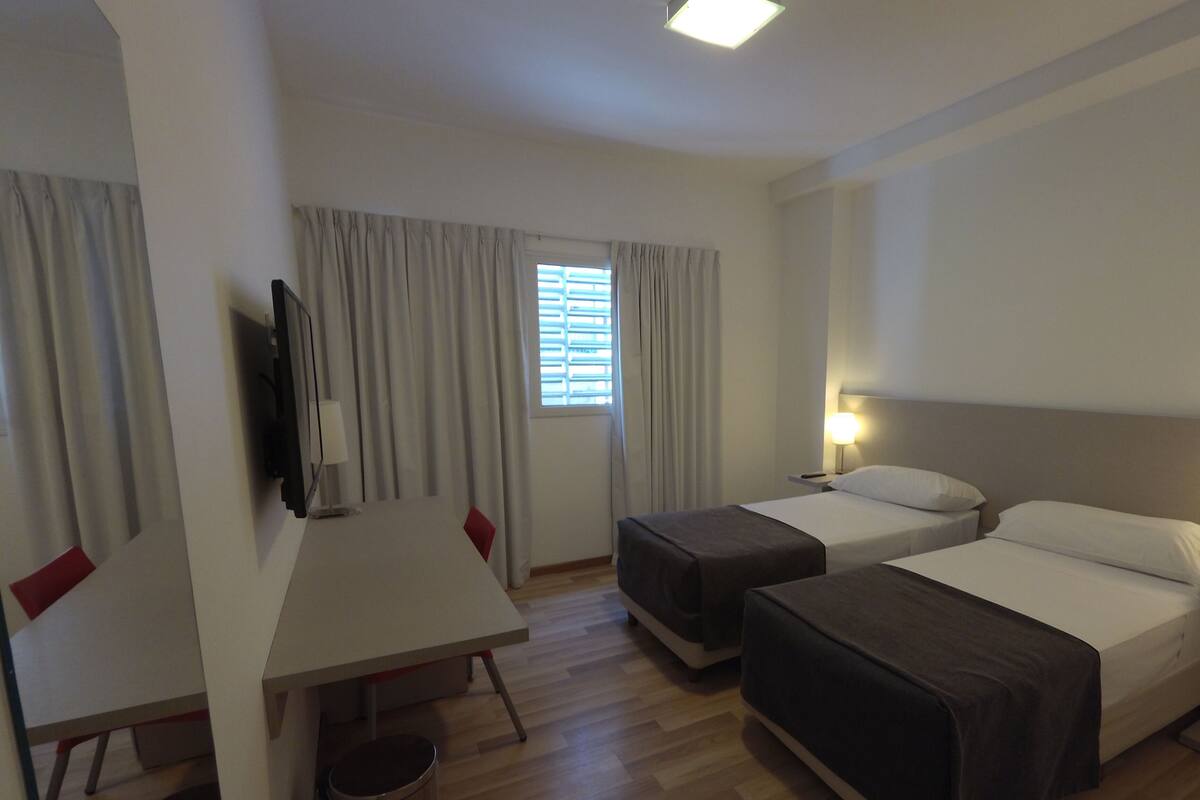 A hotel room with two single beds, a desk, a chair, a TV, and a window with curtains.
