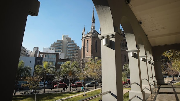 Vista de un edificio con una torre y aguja destacadas, rodeado de edificios modernos y un estacionamiento con automóviles.