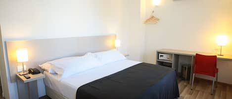 A hotel room with a bed, bedside tables, a desk, a chair, and an air conditioning unit.