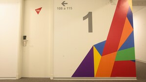 Un pasillo con una puerta marcada '108' y '1', una flecha direccional y un diseño geométrico de colores en la pared.