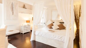 A bedroom with a canopy bed, bedside tables, a nightstand, and a wall-mounted shelf with decorative items.