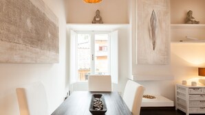 A dining area with a dark wooden table, white chairs, and a bowl of nuts. There is a large painting on the wall, a Buddha statue, and a shelf with decorative items.
