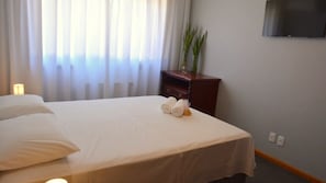 A hotel room with a bed, a nightstand, a lamp, a TV on the wall, and a window with curtains. - Locar-in Gramado - Aconchego Coração de Gramado (Gramado)