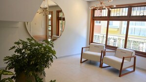 Une pièce spacieuse avec un grand miroir, deux fauteuils blancs, une plante en pot et de grandes fenêtres donnant sur un balcon.
