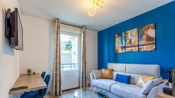 A modern living room with a white sofa, blue cushions, and a glass coffee table. A flat-screen TV is mounted on the wall, and there is a window with curtains. - "Lumières", downtown Lyon, University of Medicine/Lyon 3, metro 200 m away, wifi (Lyon)