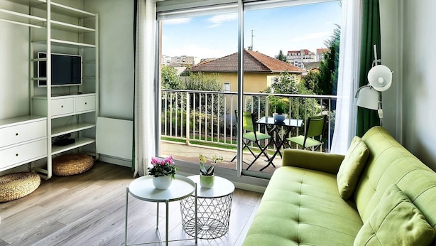 Ein modernes Wohnzimmer mit einem grünen Sofa, einem Fernseher auf einem weißen Regal und einem Balkon mit Tisch und Stühlen.