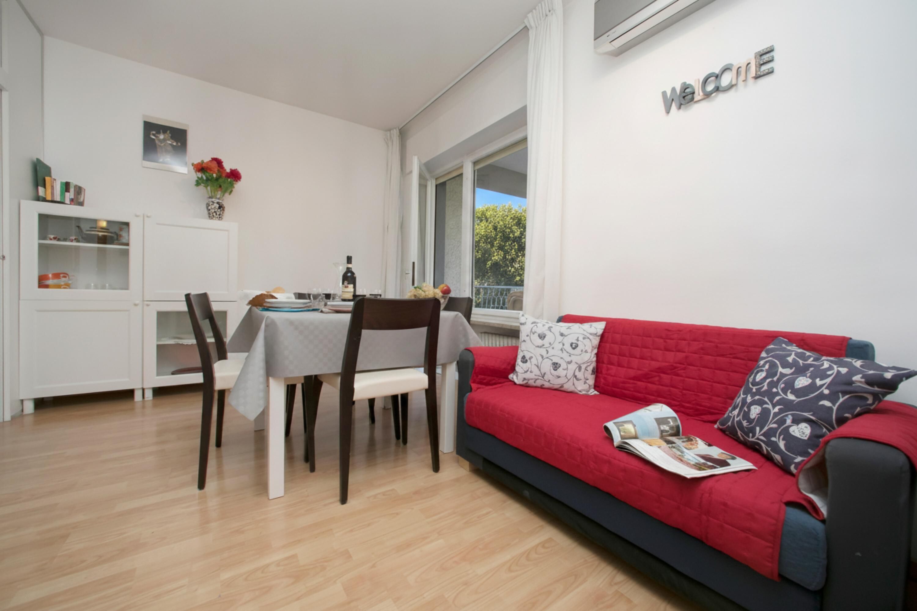 Een moderne woonkamer met een rode bank, een eettafel met stoelen, een keukenhoek en een 'Welcome'-bord aan de muur.