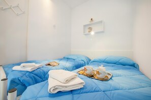 A hotel room with a blue bedspread, a white wall, a shelf with decorative items, and a tray with a teapot and cups. - Riva Lago e Spiaggia Apartment (Riva del Garda)