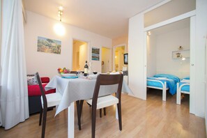 A dining area with a table set for two, a red sofa, and a bed with blue bedding in the adjacent room.