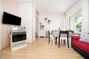 Ett modernt vardagsrum med en platt-TV, ett matbord med stolar, en soffa och en köksdel med mikrovågsugn.