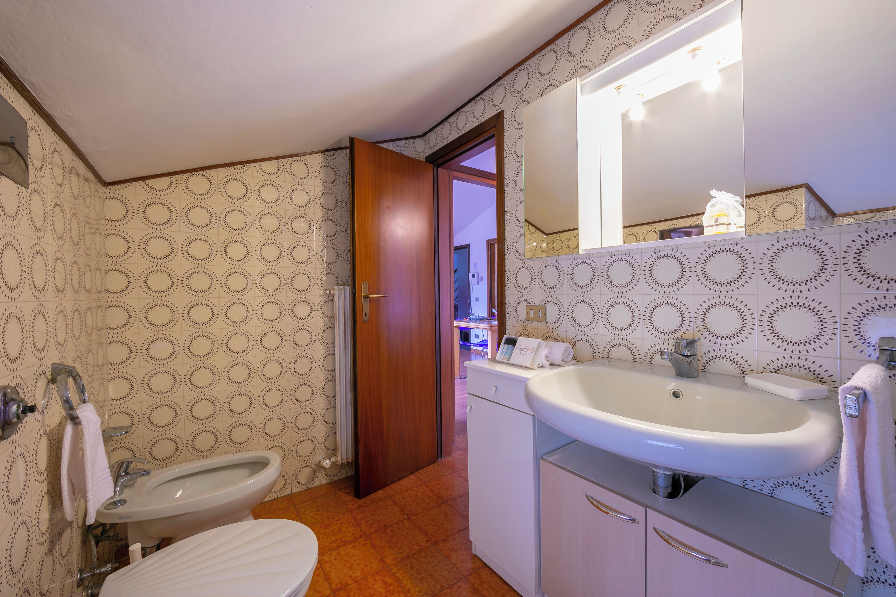 A bathroom with a toilet, bidet, sink, and mirror. There is a door leading to another room.