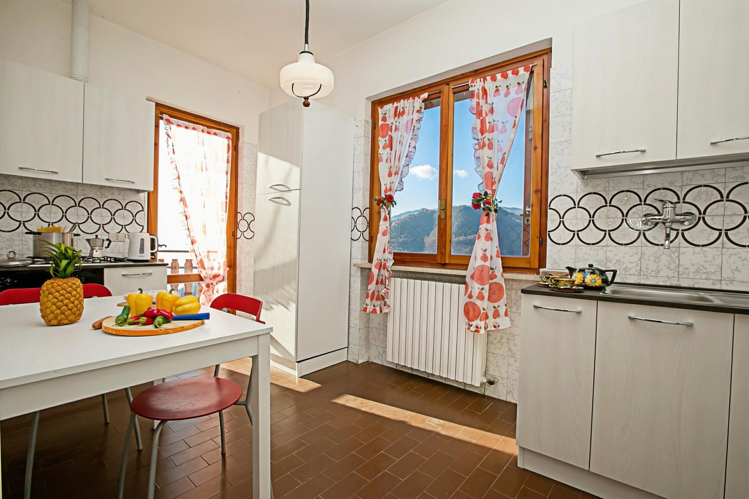 A kitchen with white cabinets, a dining table with red chairs, a fruit bowl, and a window with curtains.