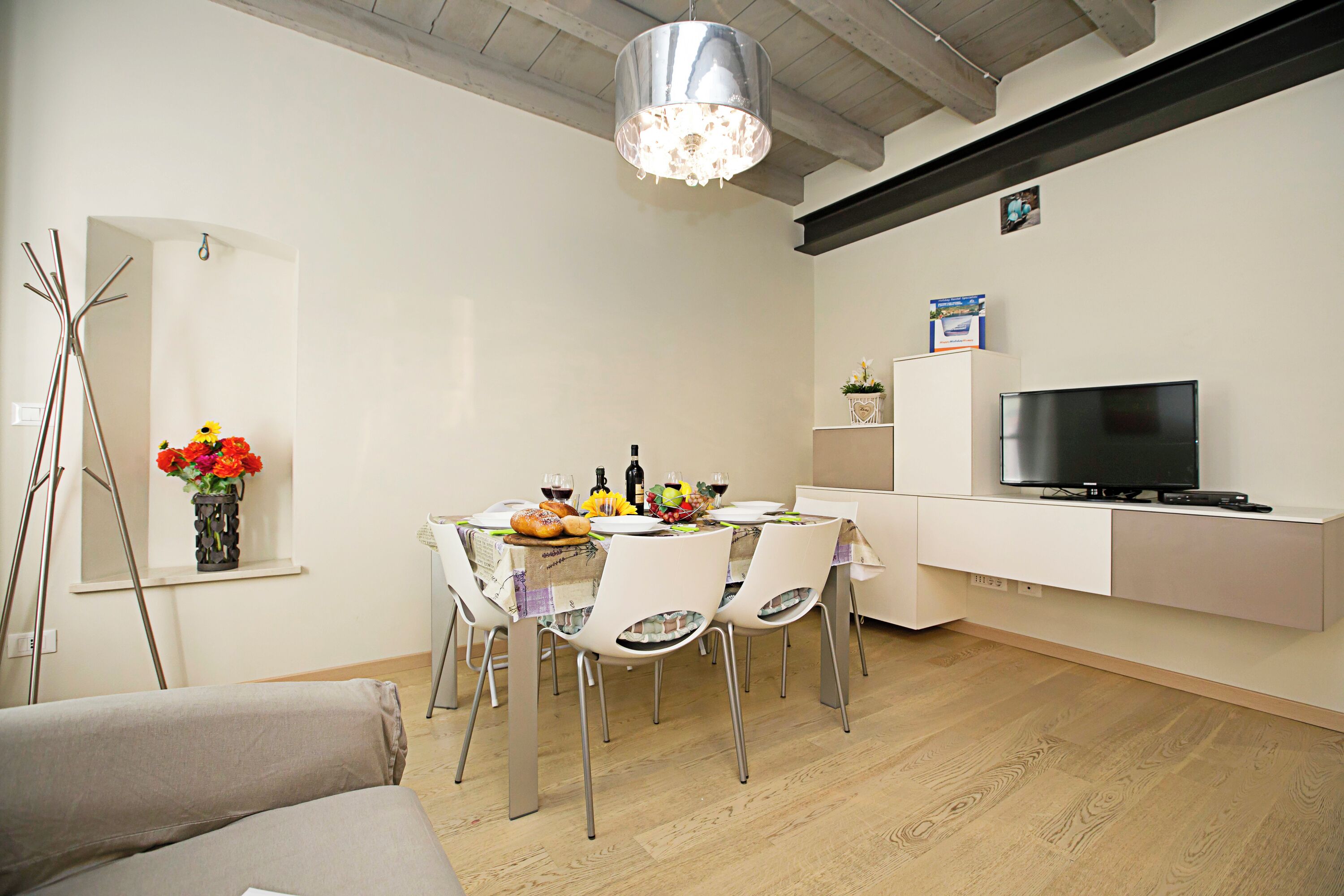 A dining area with a table set for four, a television on a stand, and a vase of flowers on a shelf.
