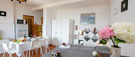 Een moderne hotelkamer met een zithoek, een bed met een grijze plaid, witte stoelen, een houten tafel en een kroonluchter.