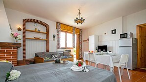 A bedroom with a bed, a desk, a chair, a dining table, and a kitchen area with appliances.