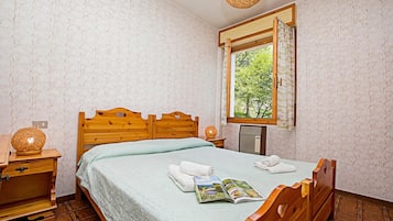 A bedroom with a wooden bed, a bedside table with a lamp, a window with a view of trees, and a patterned wallpaper.
