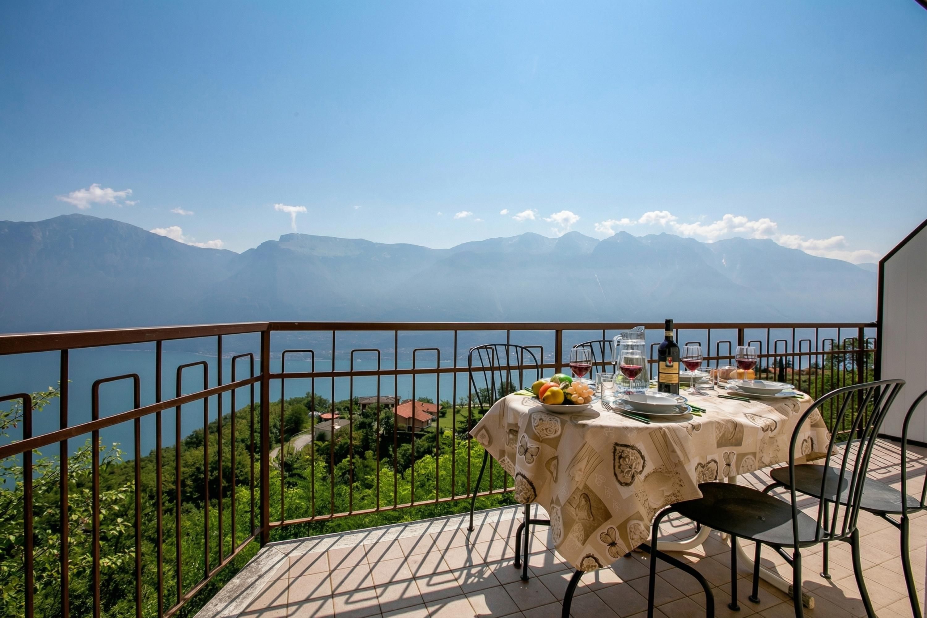 A terrace with a table set for dining, overlooking a mountainous landscape and a body of water.